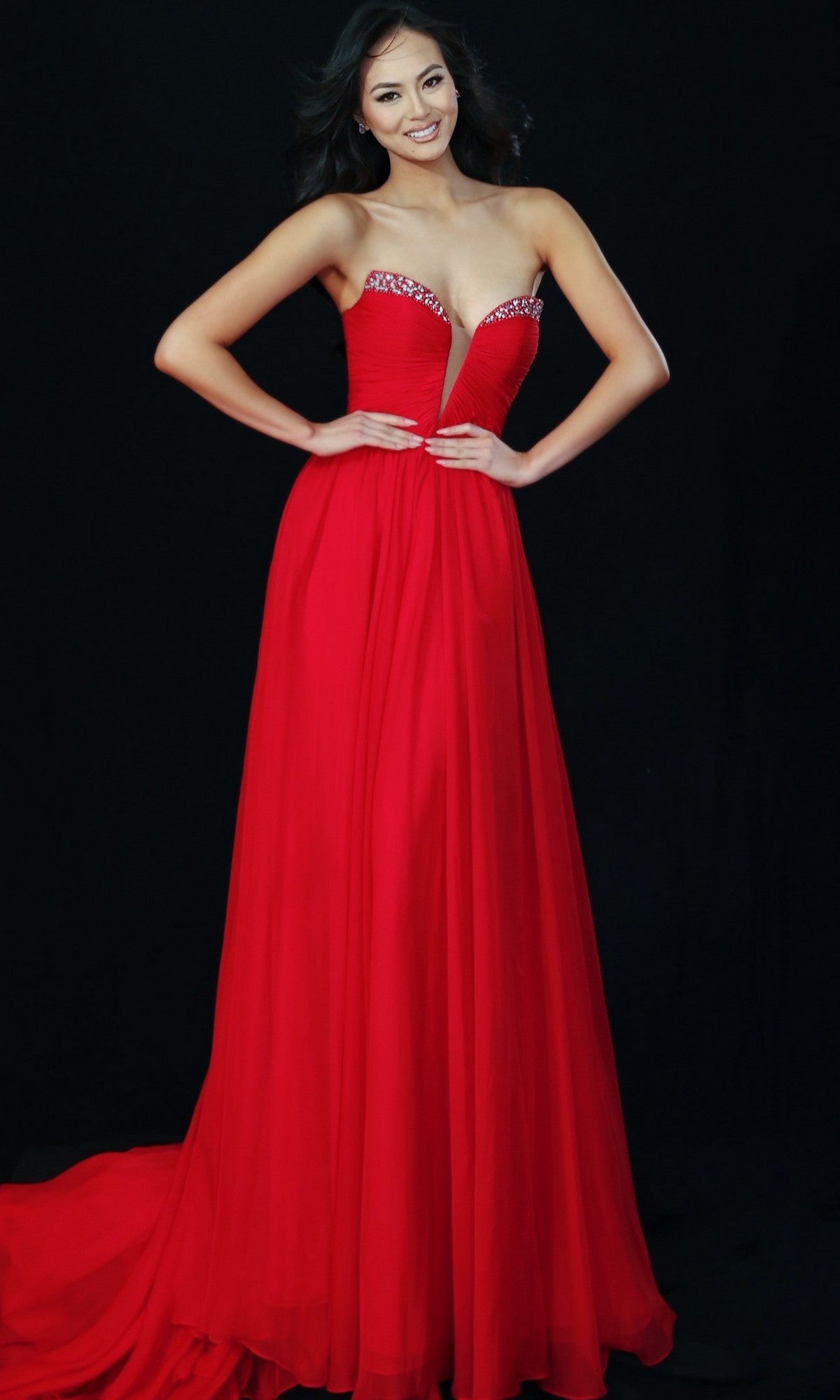A woman poses against a black background in the Jovani 49422 long prom dress, a strapless red gown with jeweled details along the sweetheart neckline and a flowing floor-length skirt.